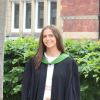 This picture shows a woman standing in front of a red brick building and a hedge. She is wearing black graduation robes with a green collar. She is smiling.