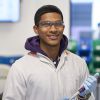 Image of a male undergraduate student standing in a laboratory. He is wearing a white lab coat with a yellow collar over a purple hoody. He is also wearing blue gloves and holding a piece of equipment. He is looking directly at the camera and smiling.