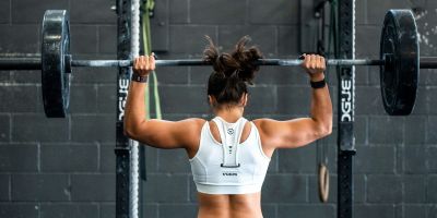 A woman lifts weight whilst facing away from the camera. She wears a white cropped vest.