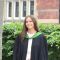 This picture shows a woman standing in front of a red brick building and a hedge. She is wearing black graduation robes with a green collar. She is smiling.