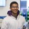Image of a male undergraduate student standing in a laboratory. He is wearing a white lab coat with a yellow collar over a purple hoody. He is also wearing blue gloves and holding a piece of equipment. He is looking directly at the camera and smiling.